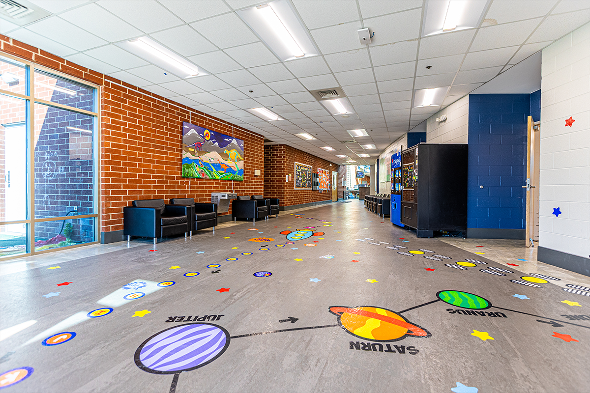 View from a lobby area down a long hallway. The floor has decals of planets, letters, stars, footprints, and a rocket ship. There are two vending machines against one wall, and black armchairs along both walls.