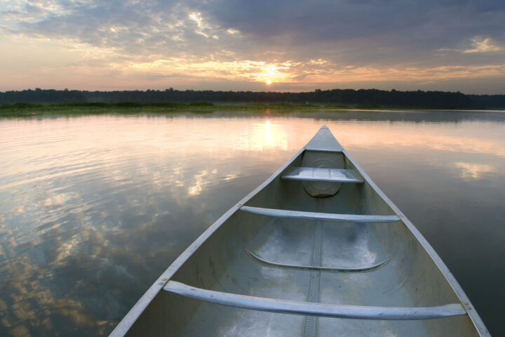 A lovely sunset viewed from the perspective of someone sitting in an aluminum canoe on a calm body of water. The bow of the canoe points toward the pink sun as it sets behind blue-gray clouds. The sunset is reflected in the still water, and trees are visible on the far shore.