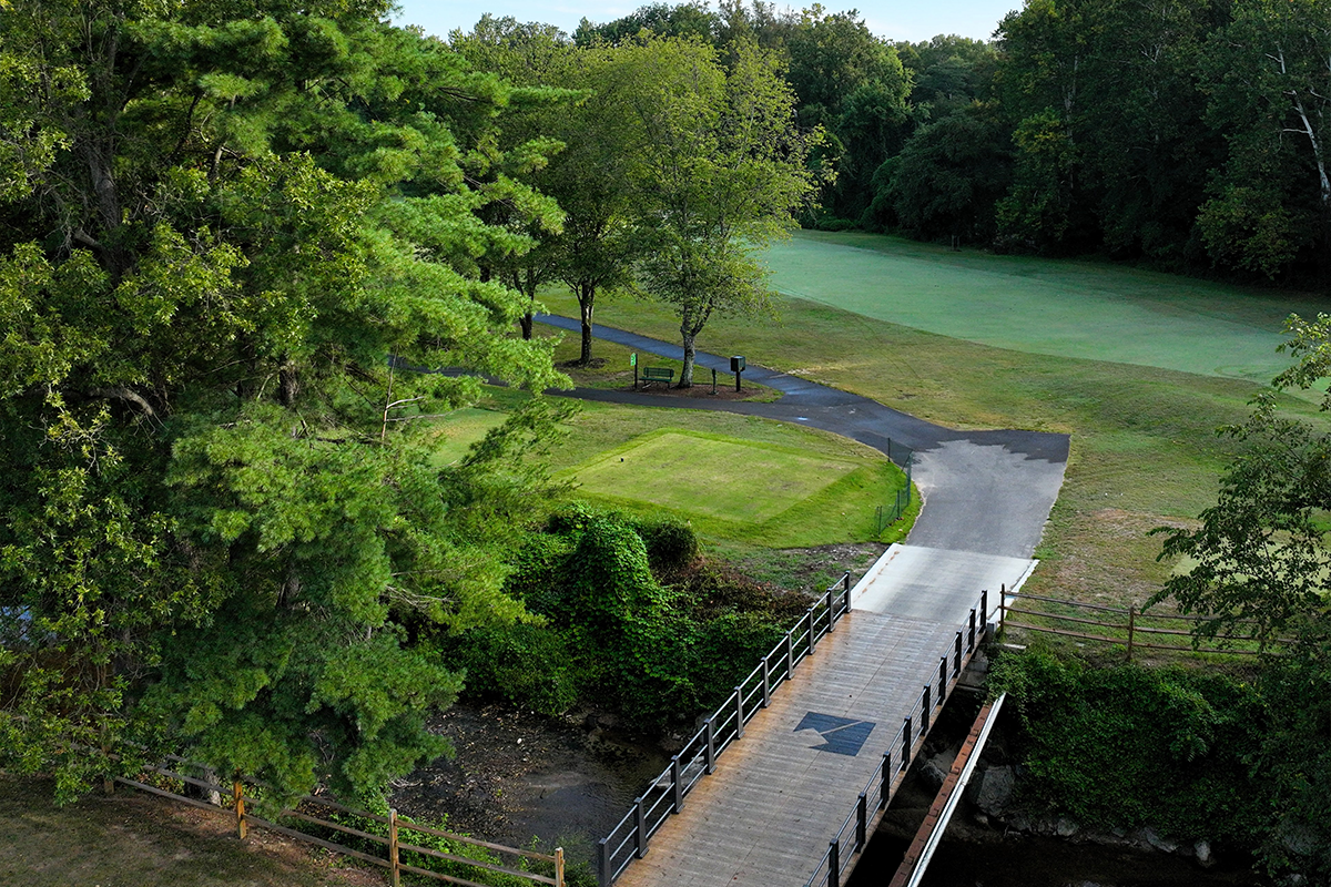 Aerial view of a wooden bridge over a creek leading to a green fairway on a golf course, flanked by a wooded area. The bridge connects to a forking asphalt path. The bridge has a black Parks and Rec logo painted on it.