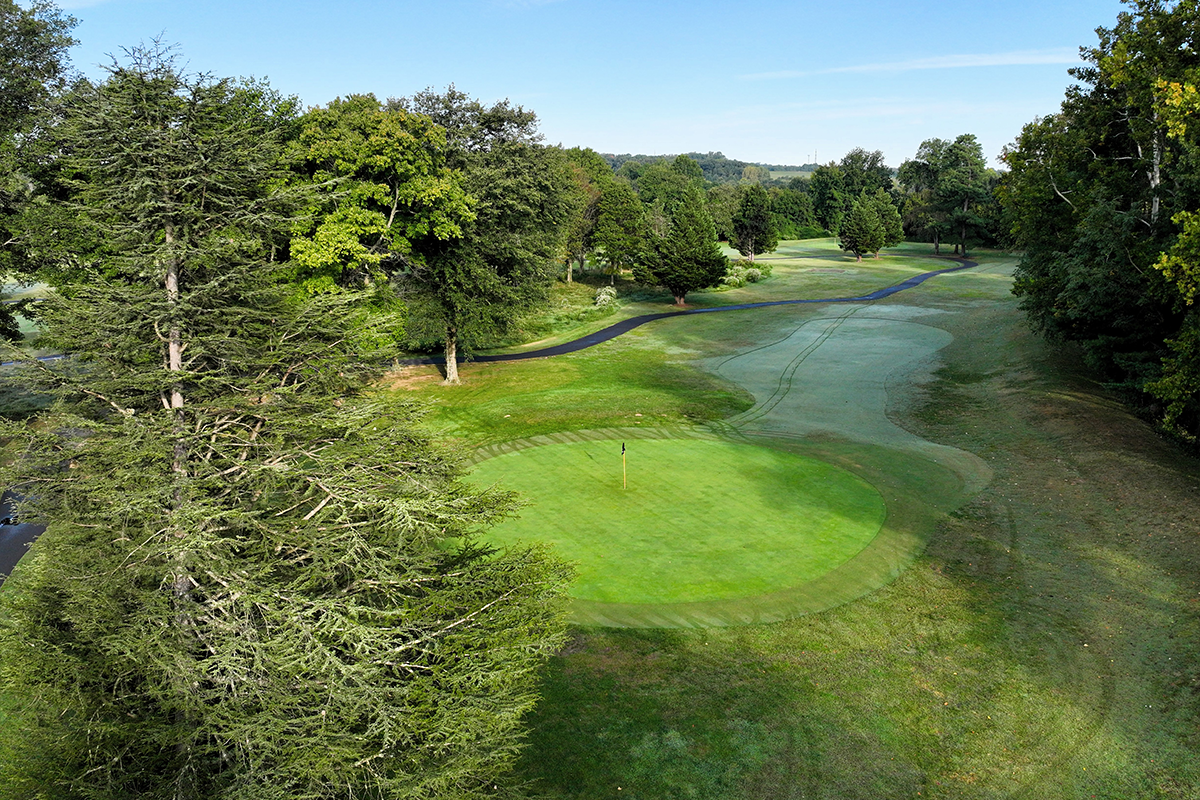 Aerial view of a hole on a golf course, with a circle of bright green grass surrounding the hole, marked by a flag. A black asphalt path winds through grass and trees beyond the hole, which is surrounded by tall trees on several sides.