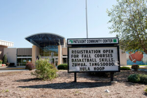 Exterior view of a sign in front of a large, beige building with a tall curved portico. Sign reads PATUXENT COMMUNITY CENTER.