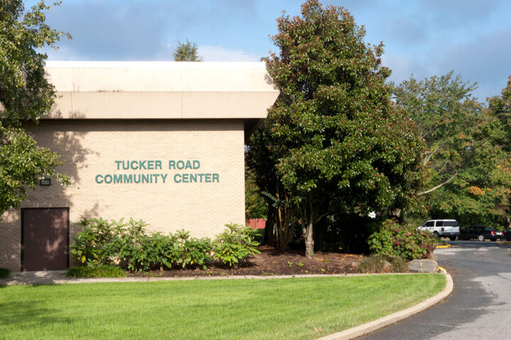 Exterior view of the corner of a beige brick building with trees around it. Green lettering on the building reads TUCKER ROAD COMMUNITY CENTER.