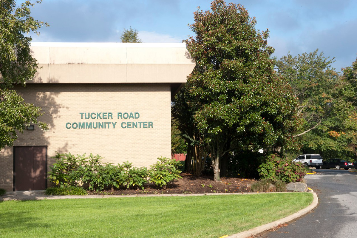 Exterior view of the corner of a beige brick building with trees around it. Green lettering on the building reads TUCKER ROAD COMMUNITY CENTER.
