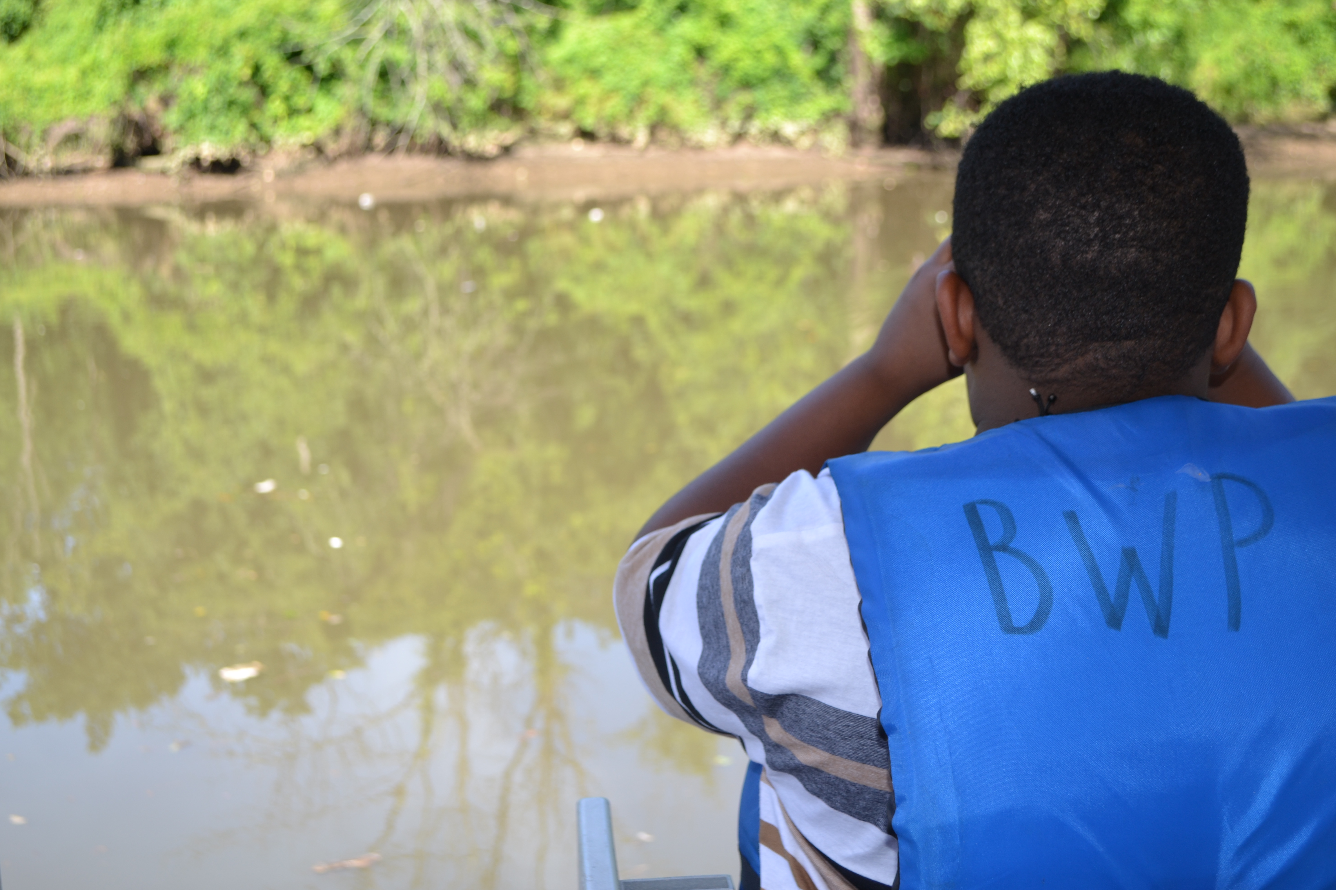 A youth looks through binoculars at a river. He is wearing a blue life vest with the letters BWP written on the back in black marker.