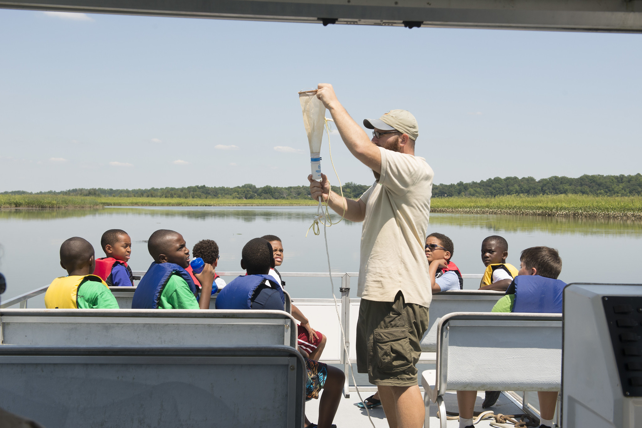 About ten kids in life vests sit on benches in the sun on a pontoon boat, all turning to look at a man in a staff t-shirt who is holding up a funnel attached to a thin rope.