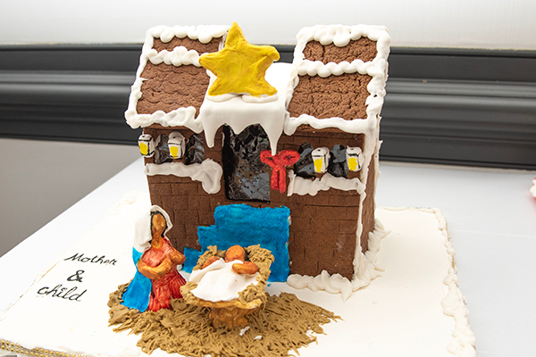 A gingerbread house with white frosting decorations, as well as a set of blue stairs leading to the door, and a giant yellow star on top of the roof. Below sits a mother kneeling over her baby sitting in a manger.