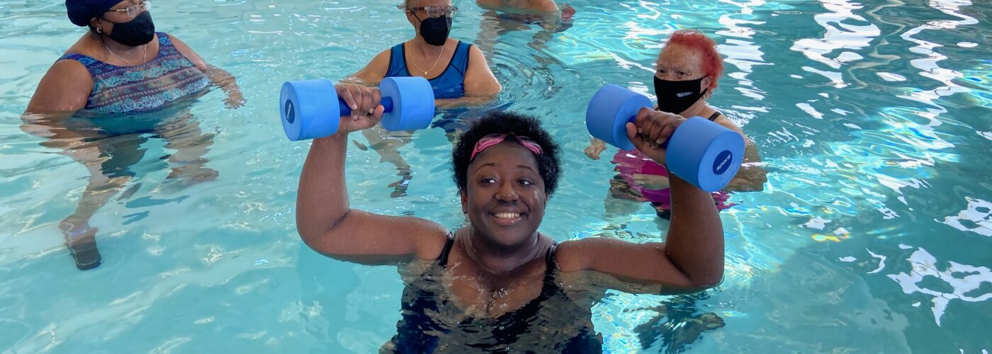 Several adults stand in an indoor pool. One is smiling and holding up blue foam weights in both hands.