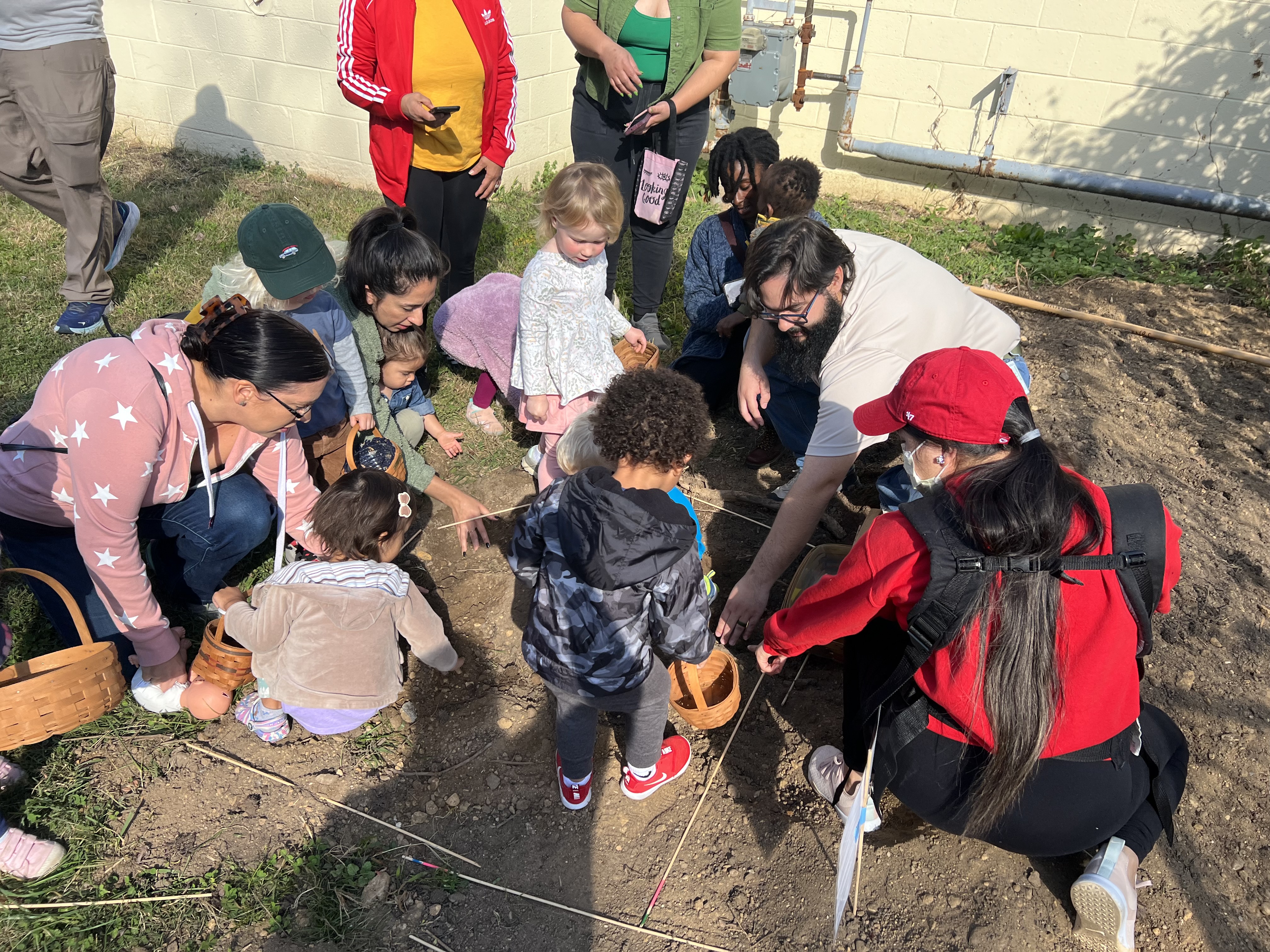 Outside, about five toddlers and their parents sit and stand on a large patch of dirt in a lawn next to a beige building. The toddlers hold small baskets.