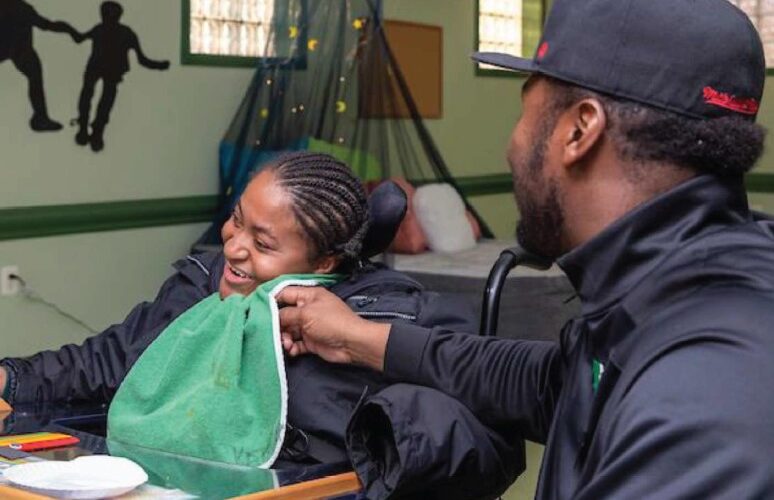 In a green room, a smiling man wearing a black staff pullover wipes the face of a smiling young woman in a wheelchair wearing a green bib over a black jacket.
