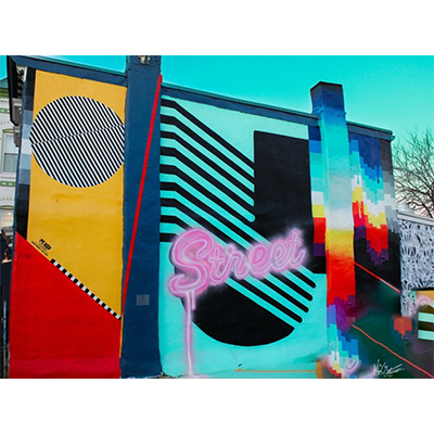 A colorful mural on the outside of a building, featuring bright colors, geometric shapes, and the cursive word "Street" styled to look like a glowing, pink neon sign.
