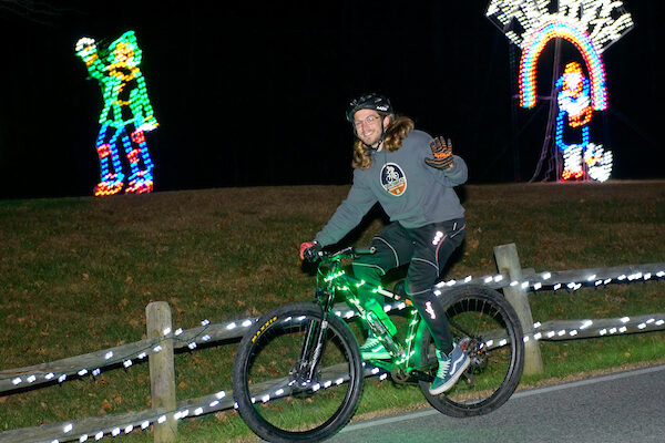 A person in a gray sweater rides a bike while waving. He is riding past holiday light decorations in the grass that are shaped like Dorothy, Toto, and the Scarecrow from The Wizard of Oz. A rainbow above Dorothy's head has the words "There's no place like home" spelled out in white lights above it.