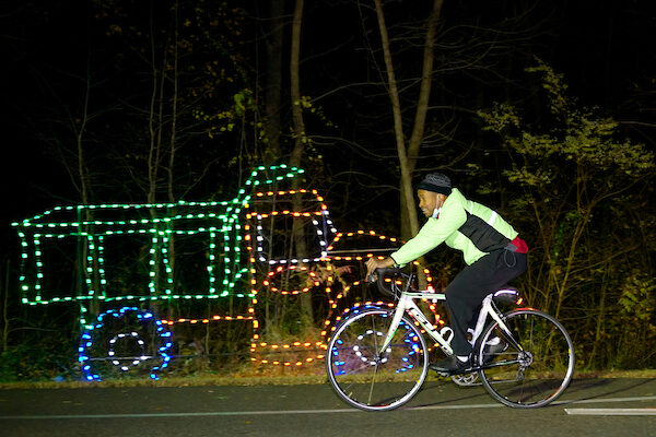A man in a green jacket rides a bike past a lighting feature in the shape of a dump truck, made of Christmas lights.