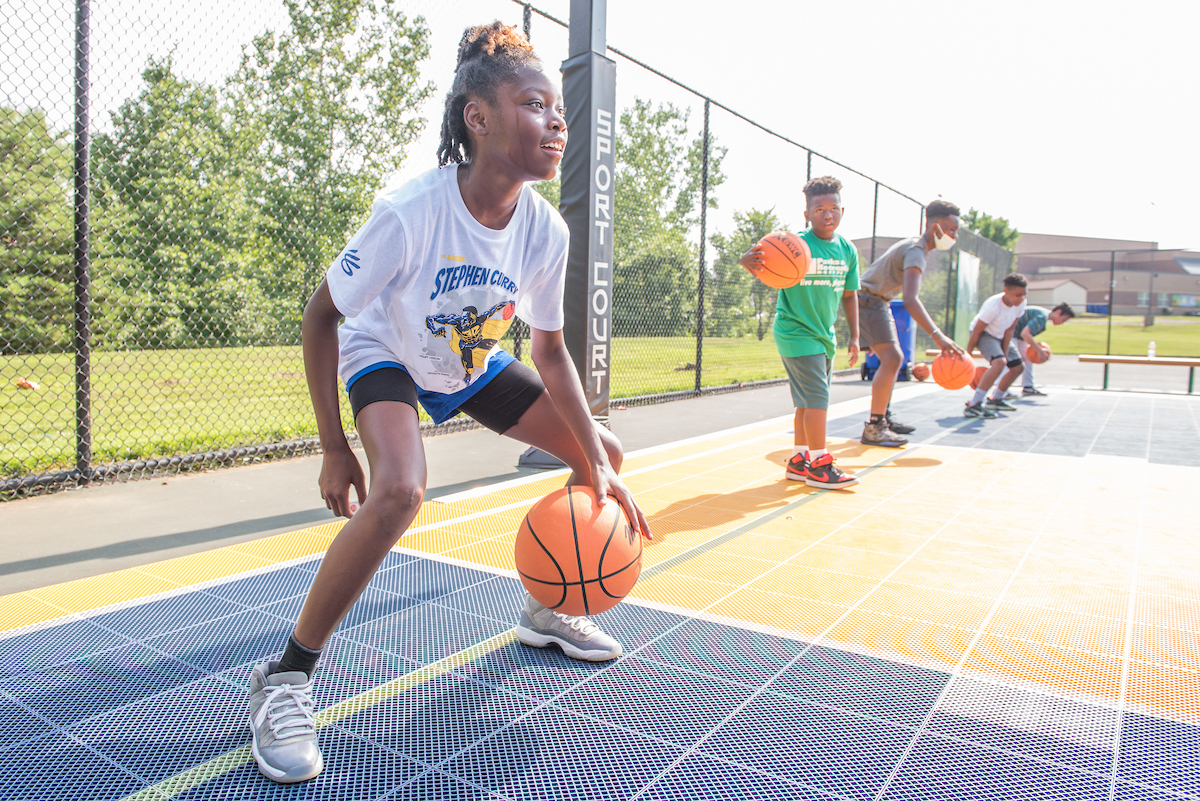 Five youths dribble basketballs on blue and yellow, permeable plastic outdoor basketball court surrounded by a chain-link fence. Grass and trees are beyond the fence. In the distance is a large, brick building.