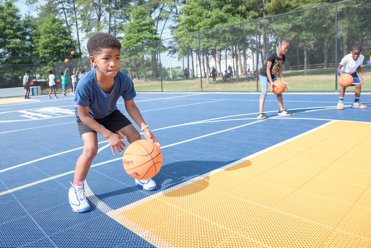 Two children dribble basketballs on a large, blue and yellow permeable plastic outdoor basketball court, surrounded by a chain link fence. Tall trees are behind the court.