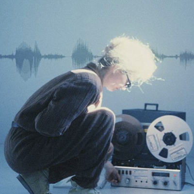 A woman in black crouches on the floor next to a reel-to-reel tape machine, adjusting one of the knobs.