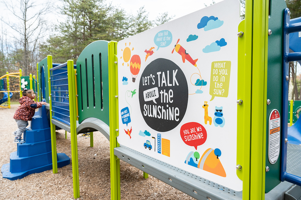 Close-up view of a long piece of play equipment at an outdoor playground, featuring raised platforms and climbing elements, with a panel featuring colorful illustrations and phrases like "Let's talk about the sunshine," and "You are my sunshine." A small child is climbing a set of blue steps at the far end.