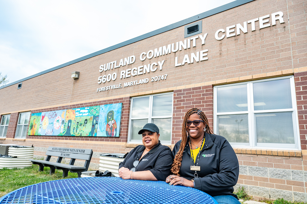 Two smiling women wearing Parks and Recreation pullovers sit at a round, blue picnic table in the grass next to a large, brick building. Letters on the building read SUITLAND COMMUNITY CENTER. Below the letters to the left is a long, colorful mural.