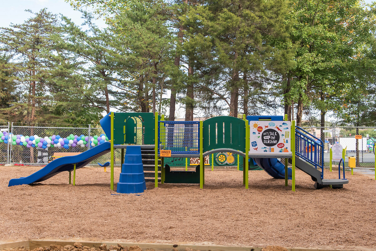 Side view of a long piece of play equipment at an outdoor playground, with slides at both ends, multiple sets of stairs, platforms, spinning features, and a curved bridge. In the background is a chain-link fence decorated with balloons.