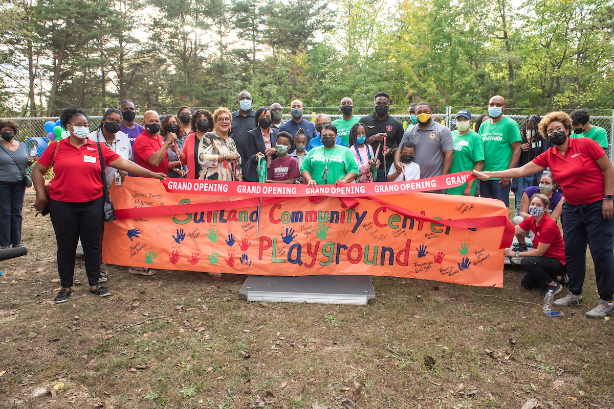 About 30 adults and a few children pose behind a large painted orange banner reading SUITLAND COMMUNITY CENTER PLAYGROUND, decorated with colorful handprints. Two staff members in red shirts hold a red ribbon that says GRAND OPENING, and four of the adults hold oversized pairs of scissors, ready to cut the ribbon.