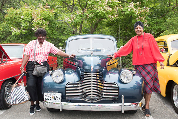 Two woman standing on either side of a 1939 Buick in a parking lot with blossoming trees in the background
