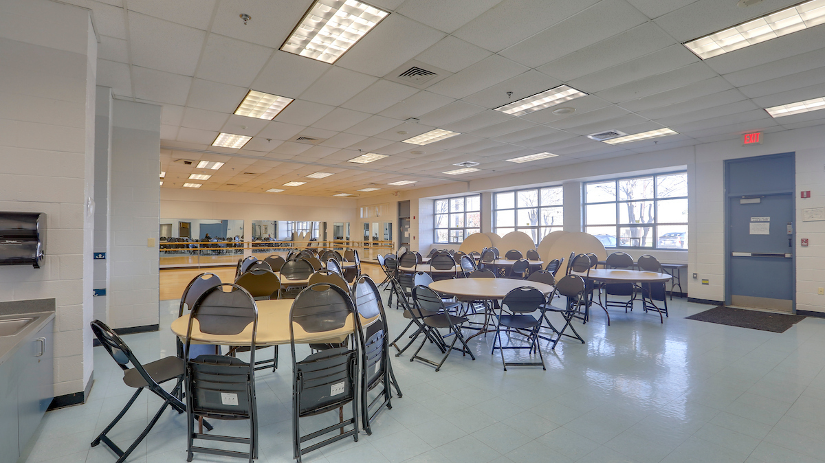 A large multipurpose room with a bank of windows along the far wall. The room contains seven round tables surrounded by folding chairs; at the far end of the room there is a dance studio with a wooden floor, mirrored walls, and barres.