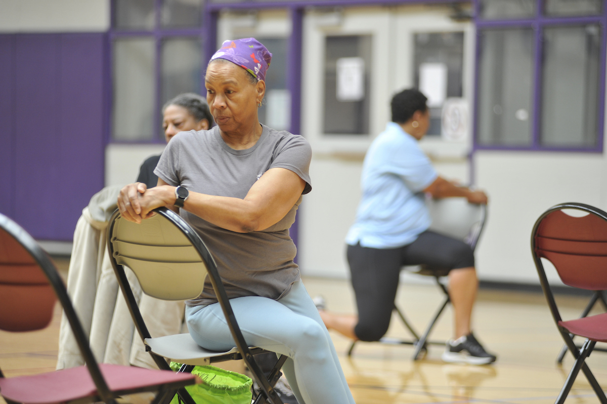 In a gymnasium with purple trim, several women do seated stretches in folding chairs. Behind them, an instructor in a light blue staff shirt demonstrates the stretch.