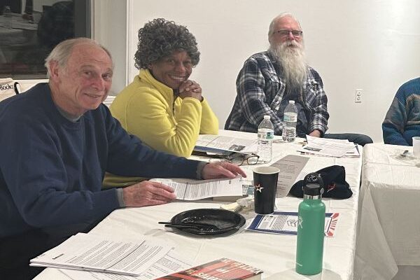 Three seniors sit at a long table covered in a white tablecloth. In front of them on the table are print-outs, books, and beverages.