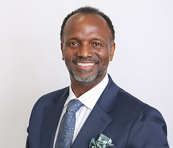 Director Stanton smiling, and looking towards the camera. He is in a blue suit, with a blue patterned tie, and is standing against a white background