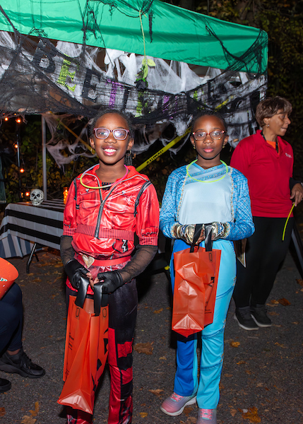 Two kids in Halloween costumes hold orange trick-or-treat bags. Behind them is a green tent decorated for Halloween with spiderweb garland and lights.