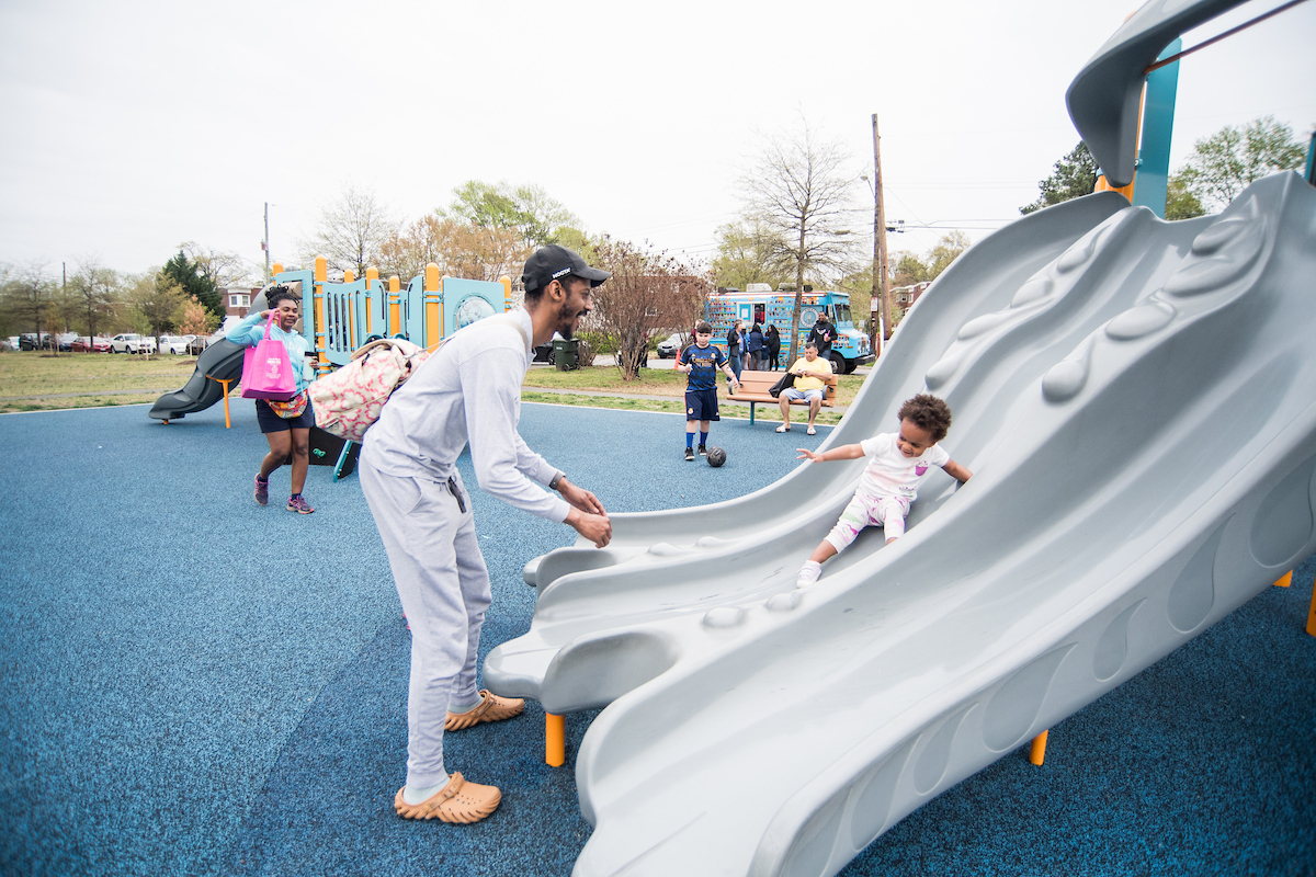 At an outdoor playground, a father stands at the bottom of a gray triple slide, preparing to catch his toddler, who is sliding down the middle track of the slide. Behind them is another play structure with more slides, platforms, and a climbing wall.
