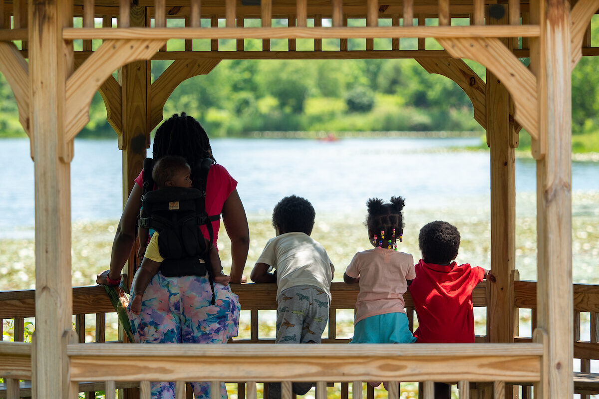 A mother and her four small children look out across the water of a lake from inside a covered wooden gazebo. The smallest child is being worn in a carrier on the mother's back.