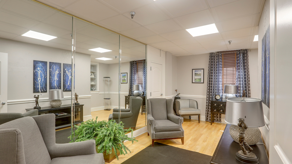 A small room with hardwood floors, one mirrored wall, several gray upholstered armchairs, and tasteful gray side tables.