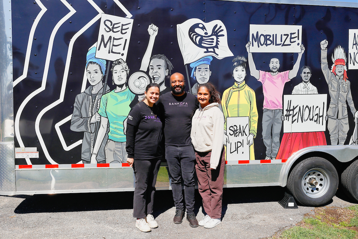 Three people wearing shirts and sweatshirts that say SANKOFA pose next to the side of the mobile museum trailer. It is painted with a large mural featuring protestors with protest signs.