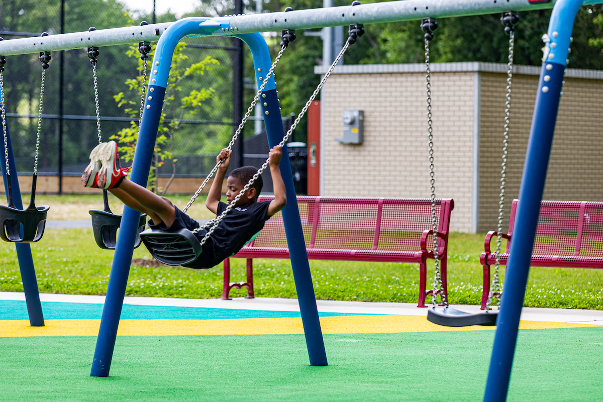 A child swings on a blue swing set at an outdoor playground. Behind it are several metal benches.