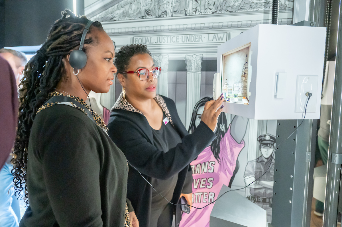 Two women interact with a small, touch-screen video monitor museum exhibit. One of them wears headphones that are connected to the exhibit. Behind them is an illustration of the Supreme Court building.
