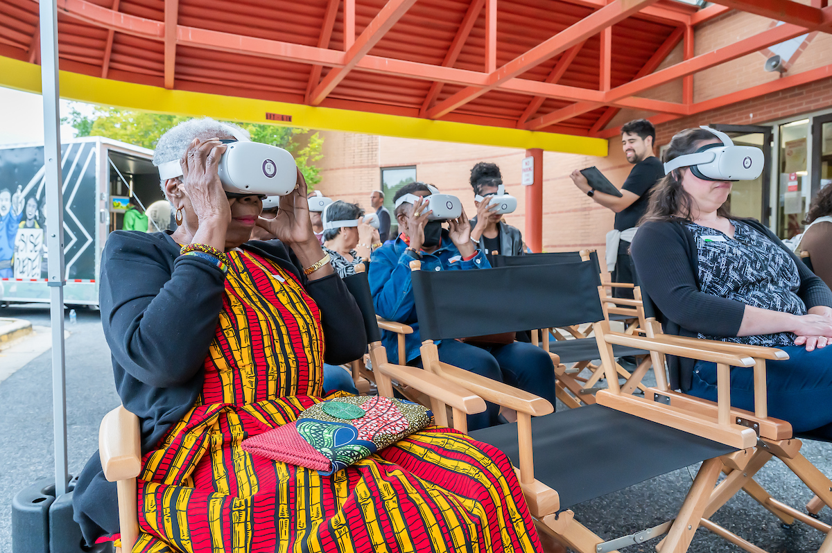 A group of people sit in black director's chairs with virtual reality headsets covering their eyes. The woman closest to the camera is wearing a bright red and yellow dress with an African print. In the background, the trailer of the Sankofa mobile museum stands open for visitors.