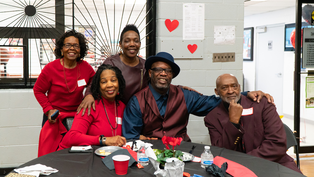 In a gymnasium decorated for Valentine's Day, five smiling adults in fancy outfits pose at a table with red flowers in the middle. They are all wearing red or burgundy outfits for the Valentine's theme.