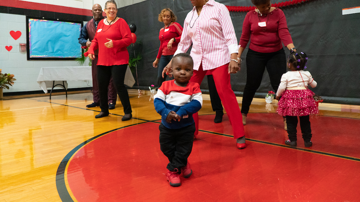 Inside a gymnasium decorated for Valentine's Day, several adults wearing red and black dance near two small toddlers, both wearing fancy outfits for the occasion.