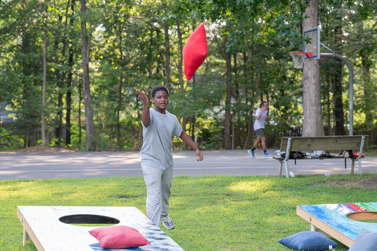 A child tosses a large, red beanbag toward an oversized game of cornhole on the grass. Behind him is an outdoor basketball court surrounded by tall trees.