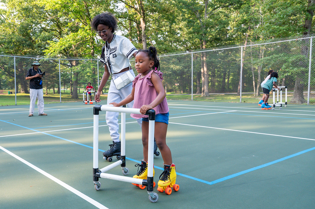 With an adult next to her, a small child holds onto a plastic support frame while roller skating on an outdoor tennis court. Behind them are other children skating with support frames. Beyond the fenced-in tennis court are tall trees, grass, and a paved path.