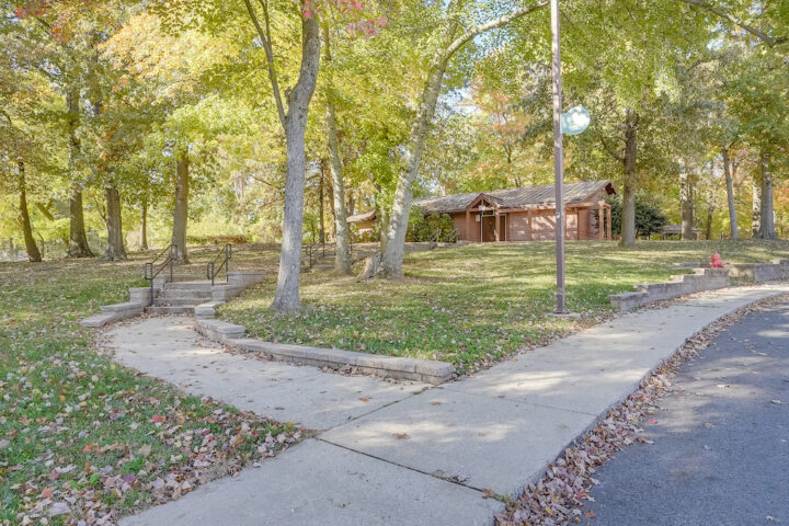 From an asphalt road, a sidewalk leads up a grassy hill, up a set of concrete steps, toward a single-story brick building surrounded by trees.