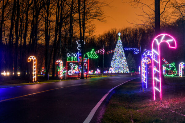 A nighttime holiday light display along a wooded road featuring large illuminated candy canes, a brightly lit Christmas tree, and colorful light sculptures including a train and other festive shapes, all glowing against an orange-tinted sky.