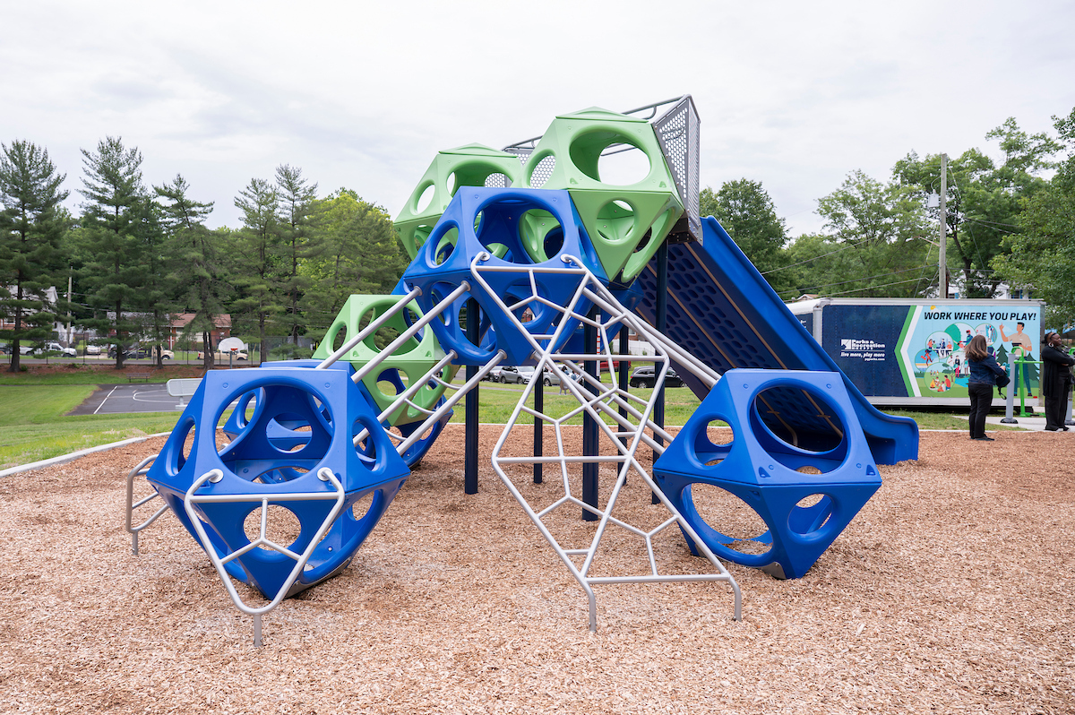 A large, blue and green climbing structure at an outdoor playground, with an extra-wide slide down one side. The structure is comprised of 3-D, hexagonal shapes with round holes in all their facets, joined into a vertical structure.