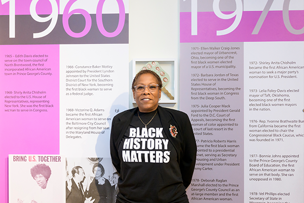A woman wearing a shirt that reads "Black Lives Matter" standing against a background covering important dates and topics from the 60s and 70s about African American culture.