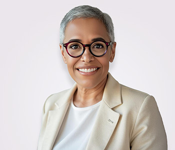 A smiling woman with short white hair wearing a beige suit jacket and red glasses.