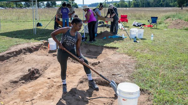 A person standing and digging in a field during the day holding a shovel next to a white bucket