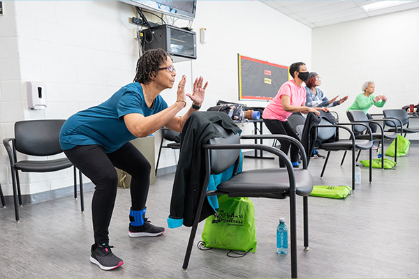 In a white room, four senior women in exercise clothes crouch behind black chairs with their arms slightly raised. There are green drawstring bags on the floor by each chair with the words "Our path to wellness" on them.