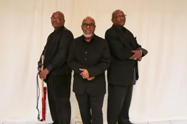 Three men wearing glasses and black suits pose together, standing shoulder-to-shoulder. One holds a red electric guitar.