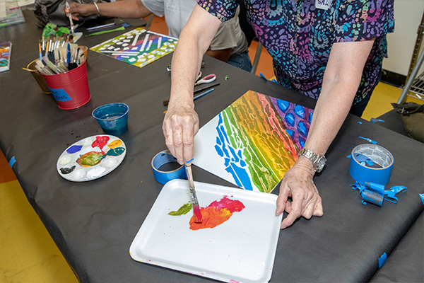 A person mixes paint with a brush in a styrofoam tray next to their partly-finished canvas, which has a colorful, abstract design painted on it.