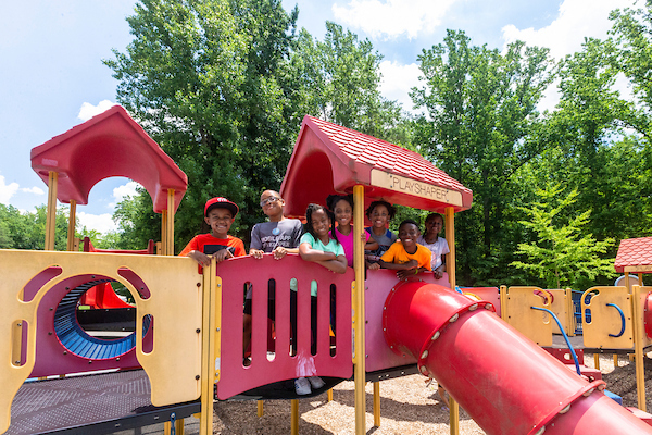 Seven kids pose on the bridge of a red playground structure. Several pathways, slides, and tunnels lead to other play areas.
